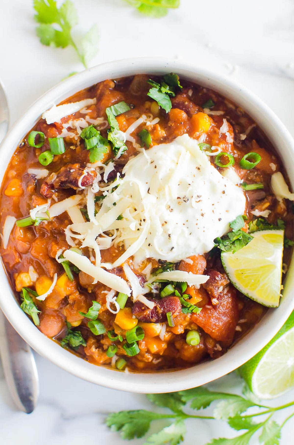 Instant Pot turkey chili with sour cream, cheese, lime and green onions in white bowl. Spoon, cilantro and lime wedge on a counter.