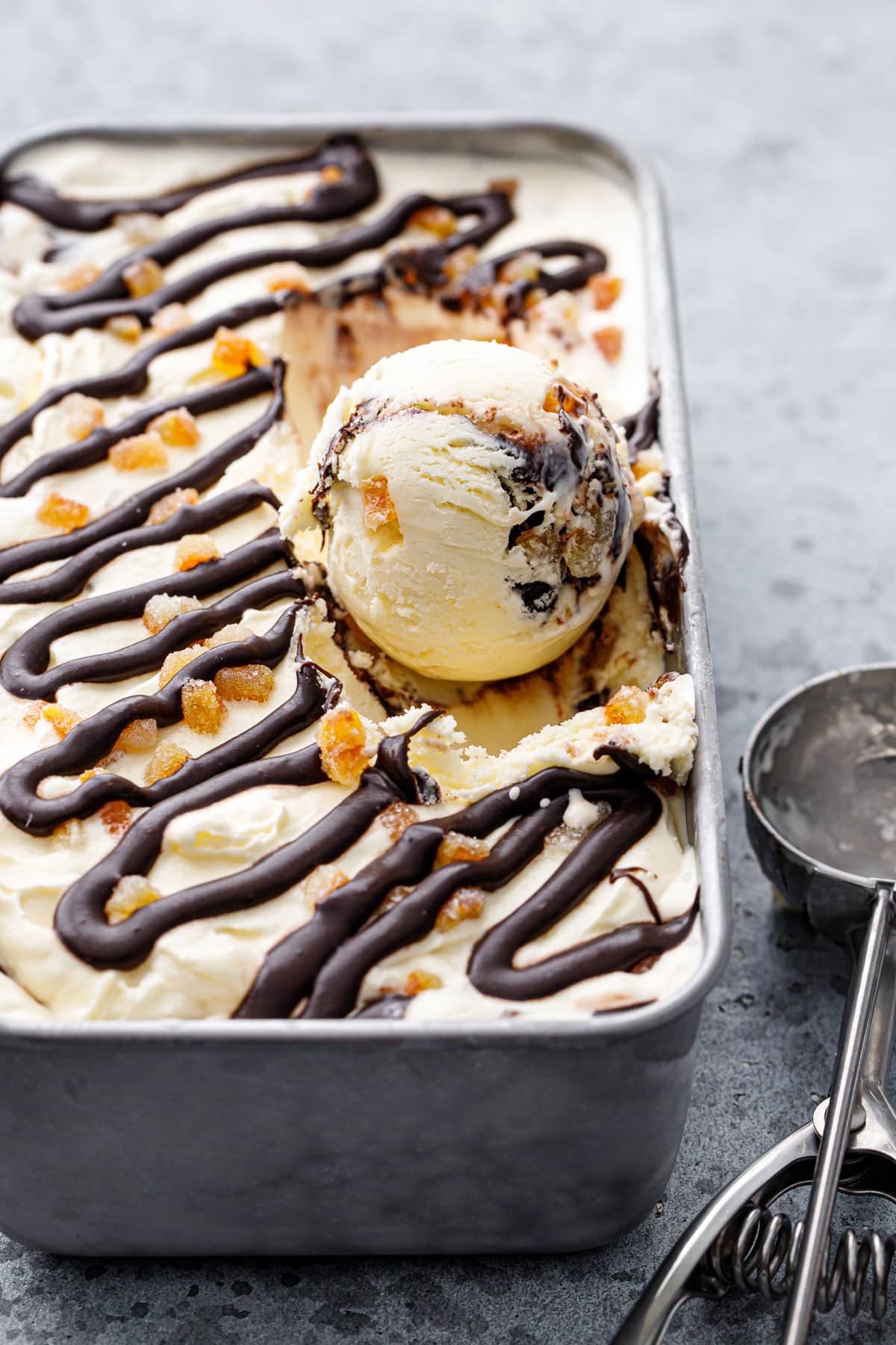 One round scoop sitting in a silver pan with Candied Orange Ice Cream with Chocolate Fudge Swirl, ice cream scoop on the side.